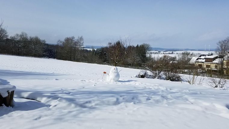 Ein verschneites Feld unter einem klaren blauen Himmel. Im Hintergrund sind Bäume und einige Gebäude sichtbar.