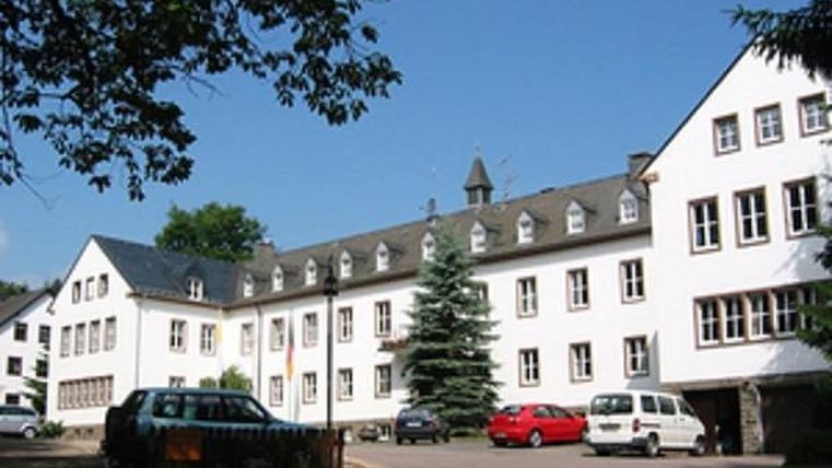 Blick auf eine großes dreistöckiges Gebäude in L-Form. Auf dem schwarzgedecktes Dach sind elf Dachgauben angebracht. Hinder dem Gebäude schaut die Spitze eines  Glockenturms hervor.
Vor dem Gebäude ist ein Parkplatz mit mehreren Autos.