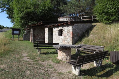 Ein alter Kalkofen aus Stein mit Holzdach, umgeben von Bänken und einem Tisch im Freien, mit Bäumen im Hintergrund.