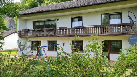 A two-story house with a balcony and large windows. In the foreground, there is a garden with a climbing frame.