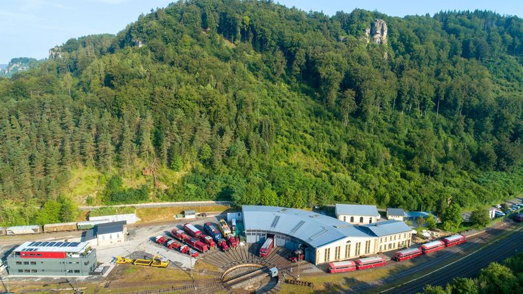 Eine beeindruckende Landschaft mit einem modernen Bahnbetriebswerk und zahlreichen Zügen. Im Hintergrund sind grüne Hügel und Bäume zu sehen.