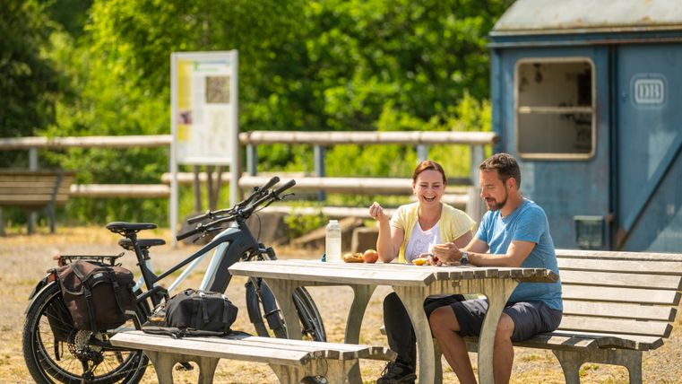 „Zwei Radfahrer sitzen auf einer Bank an einem Rastplatz entlang eines Radwegs. Ihre Fahrräder stehen neben einem Tisch. Im Hintergrund ist ein alter, blauer Lokanhänger zu sehen.“