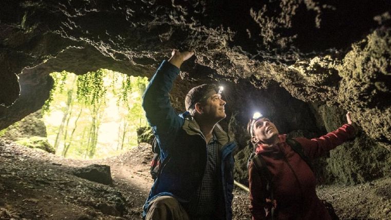 Un couple explore une grotte avec des lampes de poche. La lumière s'écoule par l'entrée et éclaire les environs.