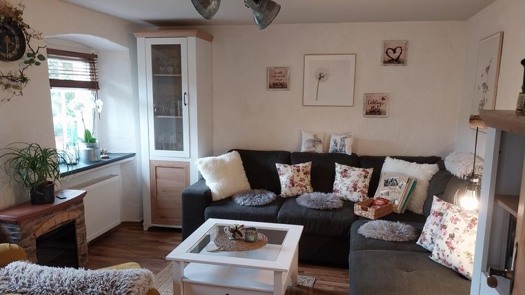 Een gezellige woonkamer met een zwarte bank en kleurrijke kussens. Aan een muur staat een glazen vitrinekast en er staan planten op de vensterbank.