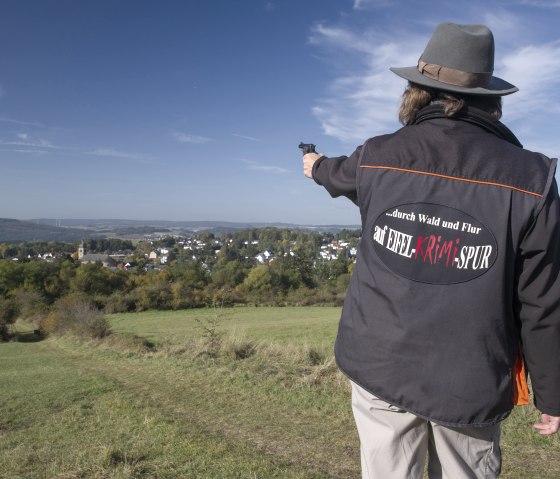 Person with hat and jacket with the inscription 'auf EIFEL KRIMI SPUR' points to a wide landscape with a village in the background., © Kappest