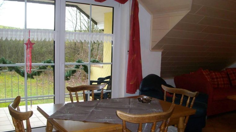 Ein gemütlicher Essbereich mit einem Holztisch und Stühlen. Im Hintergrund sind große Fenster, die auf eine grüne Landschaft blicken.