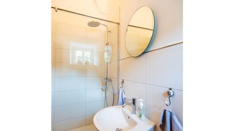 Ein modernes Badezimmer mit Dusche und einem runden Spiegel. Die Wände sind hell gefliest und es gibt einen Waschbecken mit Pflegeprodukten.