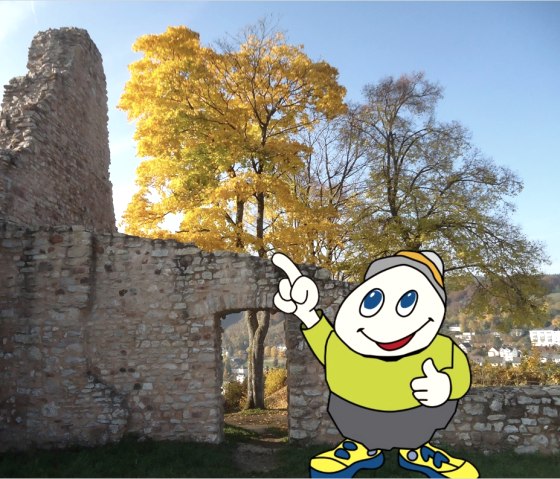 Cartoon character Willi Basalt with a green shirt, grey trousers and yellow shoes stands in front of the remains of the L&ouml;wenburg ruins and points upwards.