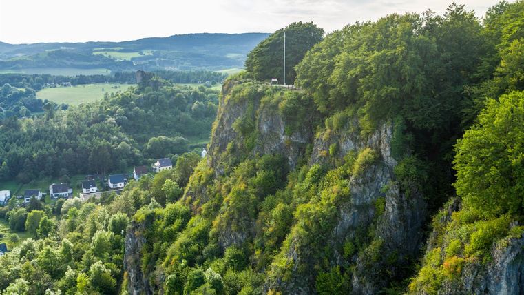 Luftaufnahme der bewaldeten Felsen der Gerolsteienr Dolomiten mit Häusern im Tal und Hügeln im Hintergrund. Ein Fahnenmast steht auf dem Felsplateau.