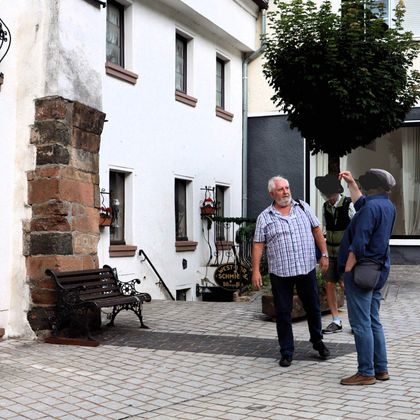 Zwei Männer stehen vor einem historischen Gebäude mit einem alten Torbogen. Einer zeigt auf etwas, während der andere zuhört.