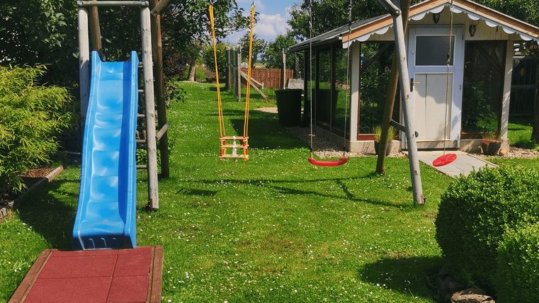 Ein schöner Spielplatz mit einer blauen Rutsche, Schaukeln und viel Grün. Im Hintergrund steht ein kleines Gartenhaus.