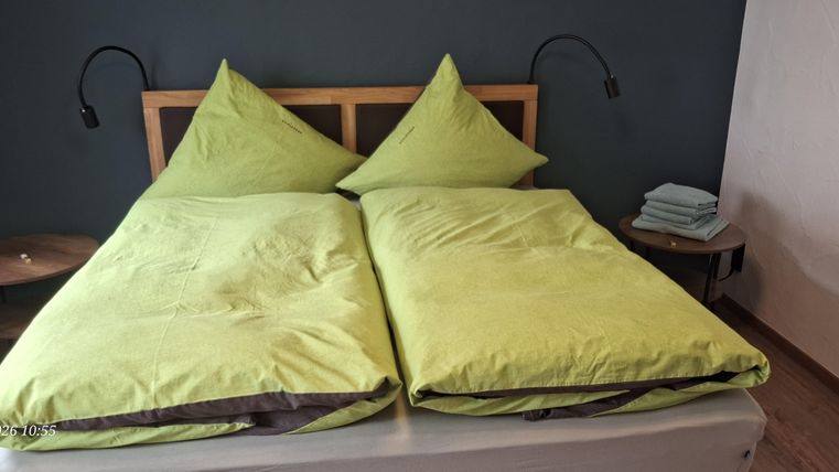Ein gemütliches Schlafzimmer mit einem Doppelbett und zwei grünen Kopfkissen. Neben dem Bett steht ein kleiner Tisch mit einer Lampe und frischen Handtüchern.