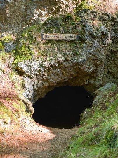 Eingang zur Borussia-Höhle, umgeben von moosbewachsenen Felsen und Gras. Ein Holzschild mit der Aufschrift "Borussia-Höhle" ist sichtbar.