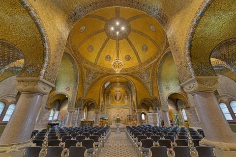 Innenansicht der Erlöserkirche in Gerolstein mit kunstvoller Mosaikdecke, Kronleuchter und Sitzreihen. Ein großes Rundfenster und Altar sind sichtbar.