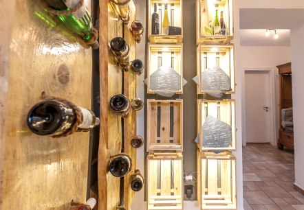 Houten planken aan een muur met wijnflessen die er met touwen aan vastzitten. Daarnaast wijnrekken van houten kisten met nog meer flessen en leisteenplaten waarop het assortiment staat geschreven.
