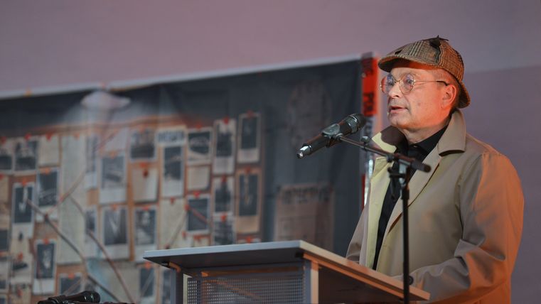 Ein Mann in Detektivkleidung steht hinter einem Podest und sprichtin ein Mikrophon.