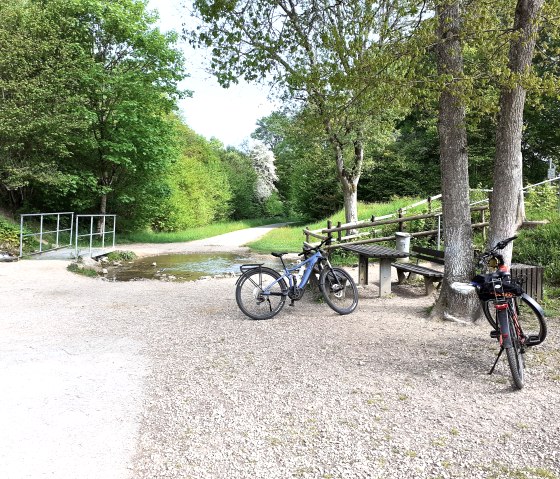Two bicycles are parked on a gravel path next to trees and a bench. A small stream flows across the path. Surrounded by green trees., &copy; Touristik GmbH Gerolsteiner Land, Ute Klinkhammer