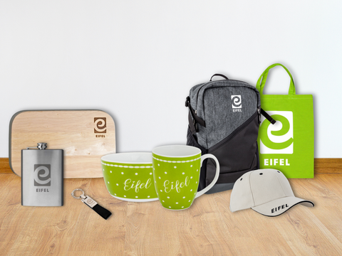 Ein Brettchen, Flachmann, Schlüsselanhänger, Schale, Tasse, Rucksack, Stoffbeutel und Basecap nebeneinander auf einem Holzboden. Alle Produkte sind aus dem Eifelshop und mit dem Schriftzug Eifel und/oder dem Logo "e" versehen.