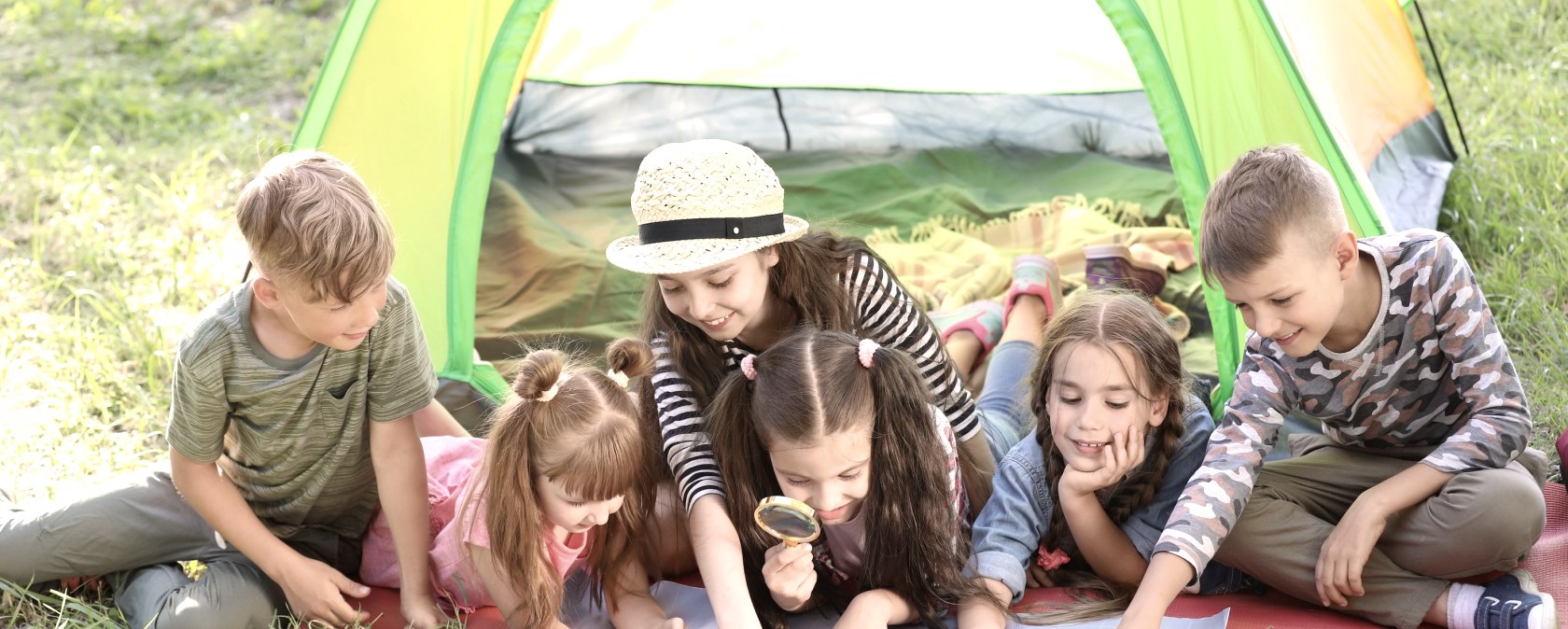 Sechs Kinder liegen und sitzen dicht beieinander vor einem bunten Zelt auf einer Wiese und schauen auf eine Landkarte, die vor ihnen liegt. Manche Kinder zeigen auf die Karte und ein Kind schaut genauer mit einer Lupe.