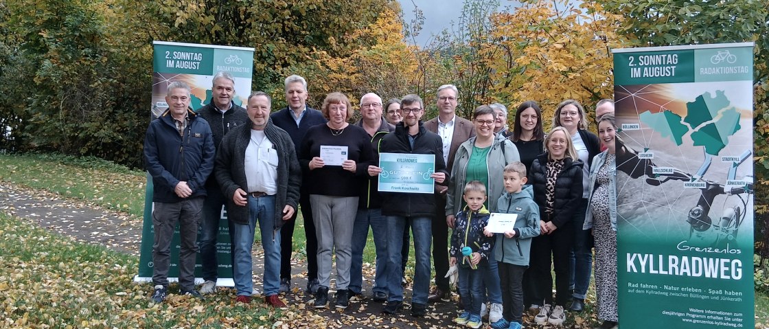 18 Personen stehen vor herbstlichen laubb&auml;umen auf einer schmalen asphaltierten Stra&szlig;e zusammen zwischen zwei Bannern f&uuml;r den Radaktionstag "Grenzenlos Kyllradweg" und l&auml;cheln in die Kamera. Drei Personen halten stolz jeweils einen Gewinn in den H&auml;nden.