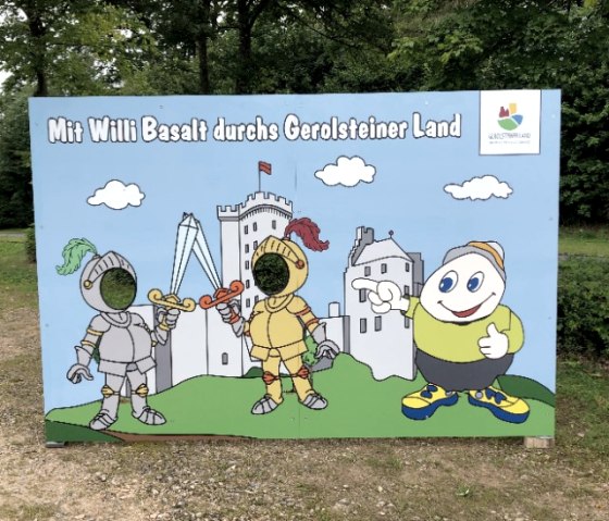 Fotowand mit Aufdruck "Mit Willi Basalt durchs Gerolsteiner Land" zeigt eine Cartoonlandschaft mit der majest&auml;tischen kasselburg in Pelm. Davor zwei Ritter, dessen Gesichter ausgeschnitten sind. Daneben das Maskottchen Willi Basalt.