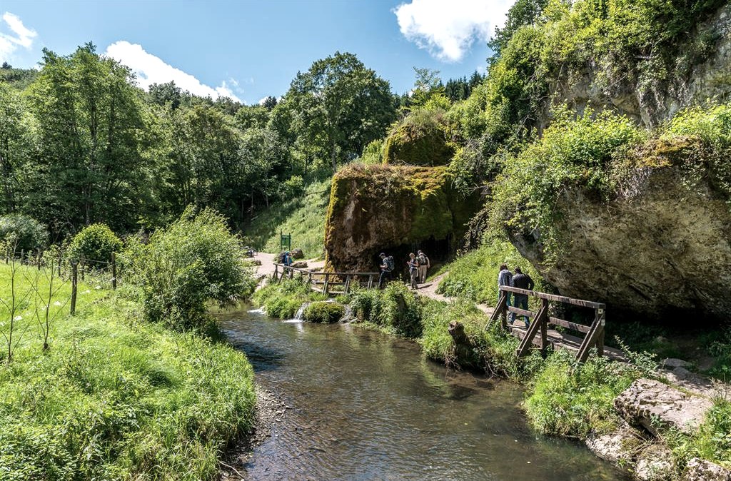Een voetpad leidt langs een kleine rivier, omgeven door weelderig groen en rotsen. Mensen lopen over een houten loopbrug., &copy; Achim Meurer