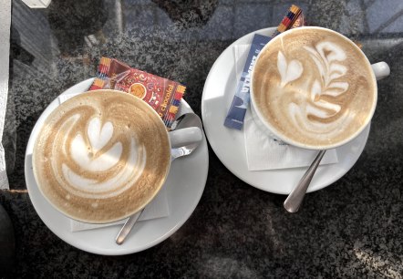 Zwei randvoll gef&uuml;llte Tassen  Cappuchino mit Milchschaumkunst auf Untertassen mit jeweils einem L&ouml;ffel, Zuckerp&auml;ckchen und einem verpackten Keks.