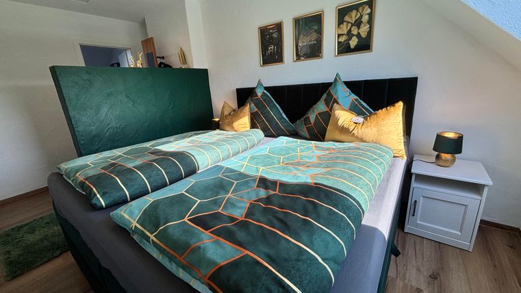 Ein gemütliches Schlafzimmer mit einem Doppelbett und dekorativen Kissen. An der Wand hängen Bilder, und es gibt einen Nachttisch mit einer Lampe.