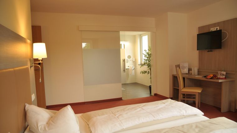 Une chambre d'hôtel moderne avec deux lits simples, un bureau et une télévision. La salle de bain et les fenêtres permettent beaucoup de lumière naturelle.