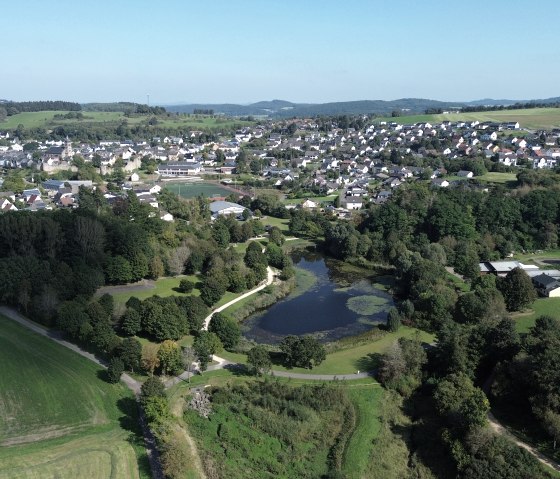 Luchtfoto van de stad Hillesheim met het kleine meer in de Bosldorfer Tälchen vallei, omgeven door bossen en velden. Op de achtergrond zijn veel huizen en heuvels te zien., © Touristik GmbH Gerolsteiner Land, Sarah Wiesen