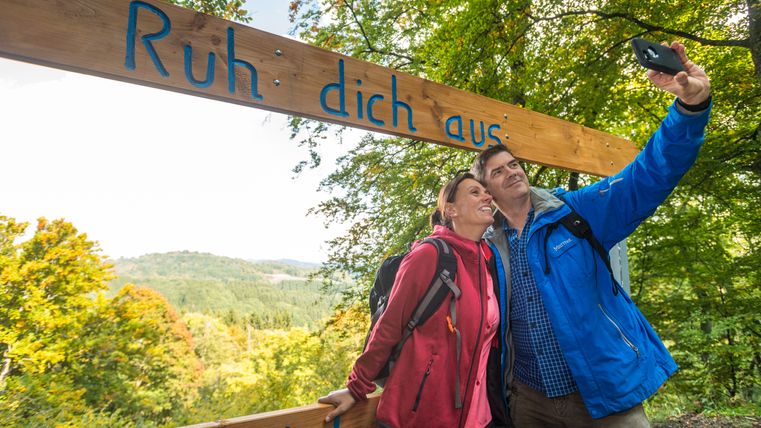 Een stel neemt een selfie onder een houten bord met de tekst 'Rust uit' in een groen boslandschap.