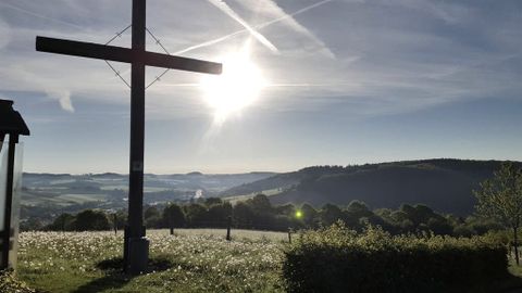 Holzkreuz auf einer Wiese mit Blick auf die hügelige Landschaft der Eifel bei Sonnenaufgang.