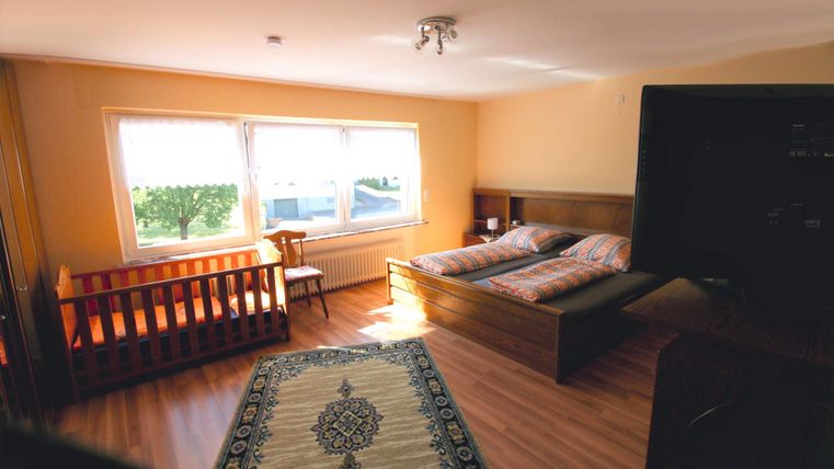 Ein helles Zimmer mit einem Doppelbett und einem Kinderbett. Große Fenster lassen viel Licht herein und eine Teppich dekoriert den Boden.
