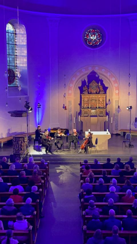 Fünf Musiker an Streichinstrumenten und Klavier musizieren vor dem Kirchenaltar. Davor die Sitzbänke voller Besucher.