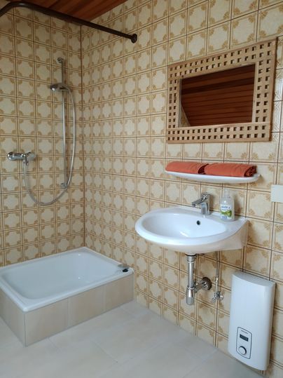 L'image montre une salle de bains avec une douche et un lavabo. Les murs sont décorés de carrelages dans un motif clair.