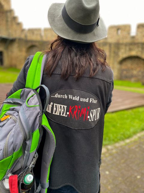 Blick auf den Rücken einer Frau, die einen Hut und eine bedruckte Jacke sowie einen grünen Rucksack über einer Schulter trägt. Vor ihr liegt die Hillesheimer Stadtmauer.