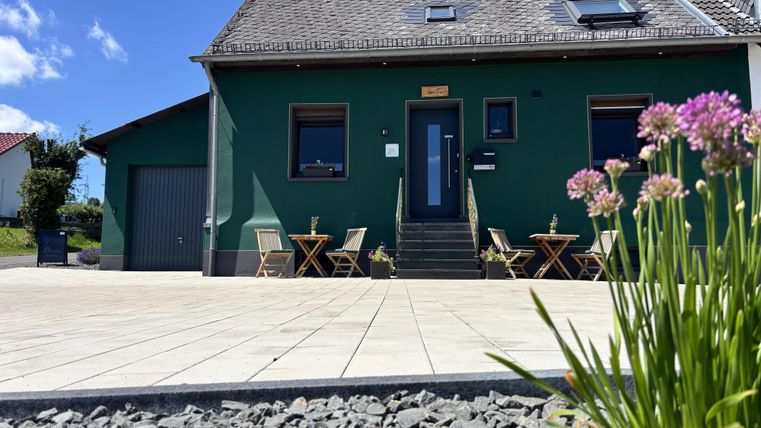 Außenfassade des Cafés und der Pension Hamony Holiday mit dunkel grüner Wandfarbe aus der Froschperspektive mit Blumen im Vorrdergrund.