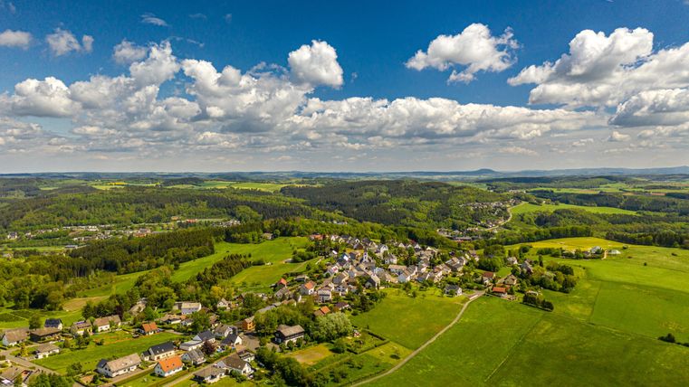 Luftaufnahme eines Dorfes in der Eifel mit grünen Feldern und bewaldeten Hügeln unter einem blauen Himmel mit Wolken.