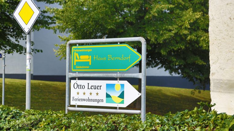 Ein Wegweiser zeigt in Richtung einer Unterkunft und Ferienwohnungen. Die Schilder sind grün und blau, umgeben von Bäumen und Gras.
