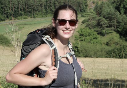 A woman in a meadow with a hiking rucksack on her back and sunglasses looks at the camera and smiles.