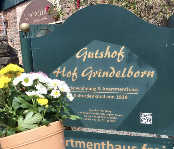Een hand houdt een grote bloempot met bloeiende bloemen erin vast voor het grote groene toegangsbord van het landgoed &quot;Hof Grindelborn&quot;