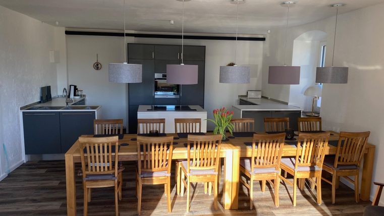 Ein modernes Esszimmer mit einem großen Holztisch und zahlreichen Stühlen. Die Küche im Hintergrund ist hell und einladend, mit eleganten Lampen über dem Tisch.