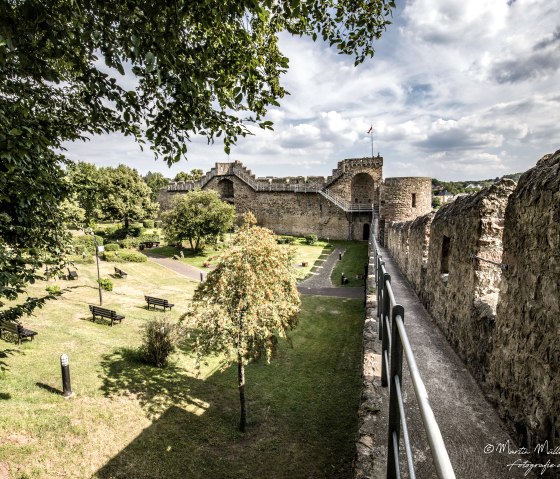 Historische stadsmuur van Hillesheim met tuin, banken en bomen. Er loopt een pad langs de muur, die is afgezet met hekwerk. Bewolkte lucht., © Martin Müller