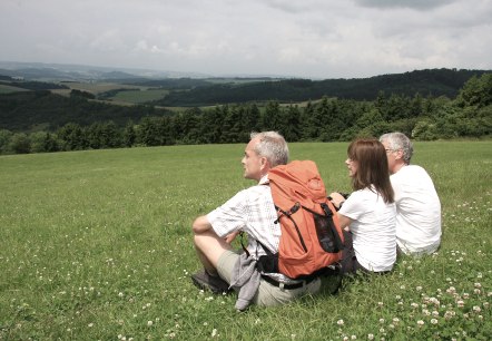 Drei Wanderer sitzen auf einer grünen Wiese und blicken in die Ferne. Einer trägt einen orangefarbenen Rucksack. Im Hintergrund sind Hügel und Wälder zu sehen., © Eifel Tourismus GmbH