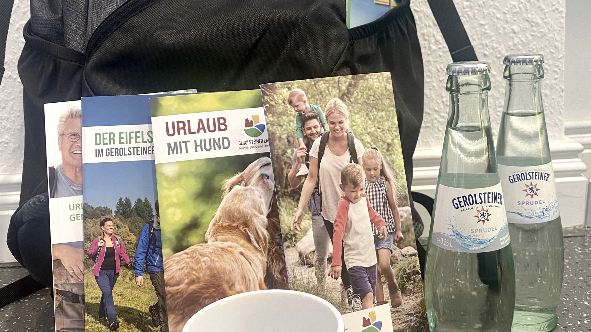 Ein Eifel-Rucksack mit mehreren touristischen Prospekten, zwei Eifel-Tassen und zwei Glasflaschen Gerolsteiner Sprudel.
