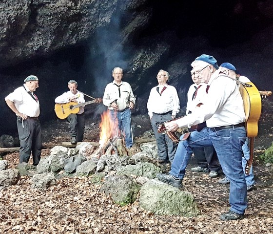 Sieben M&auml;nner mit Instrumenten stehen im Halbkreis um ein Feuer in einer H&ouml;hle.