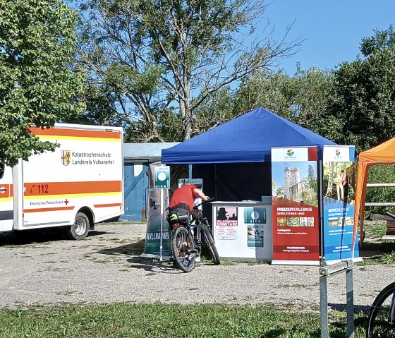 Eine Person auf einem Fahrrad hält vor einem Informationsstand auf einem Rastplatz neben einem Rettungswagen