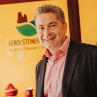 Ein schick gekleideter Herr mit Hemd und Sakko lächelt in die Kamera. Hinter ihm das Logo der Touristik GmbH Gerolsteiner Land und einige Souvenierartikel.