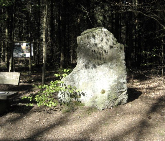 Een gedenksteen met de inscriptie 'BOB ETZEL' staat in het bos. Ernaast staat een bankje en op de achtergrond een informatiebord., &copy; Touristik GmbH Gerolsteiner Land