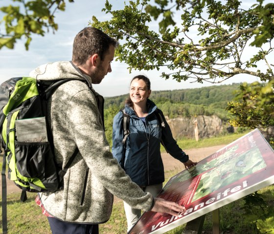 Two hikers look at an information board in the Volcanic Eifel. They are standing under a tree, with a green landscape in the background., &copy; Eifel Tourismus GmbH, Dominik Ketz